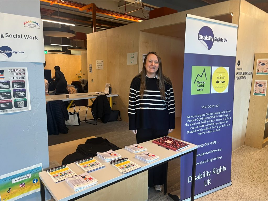 Image of Helen smiling at a Moving Social Work stall. She is standing behind a table with leaflets, next to a banner with the DR UK, GYA and Moving Social work Logos on it