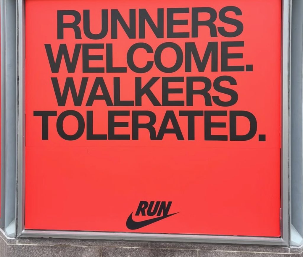 Image of a red billboard Nike advert that says Runners welcome. Walkers Tolerated. Nike logo tick underneath that says 