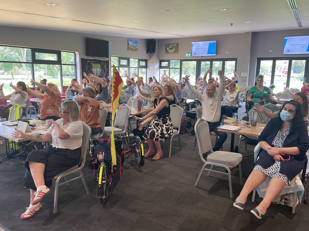 Image of a room of people sitting as an audience, taking part in seated dance as a whole group. People are smiling and enjoying themselves